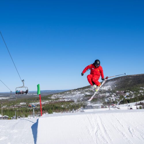 Skiers jumping in park, clear blue sky, mountains and lift in the background.