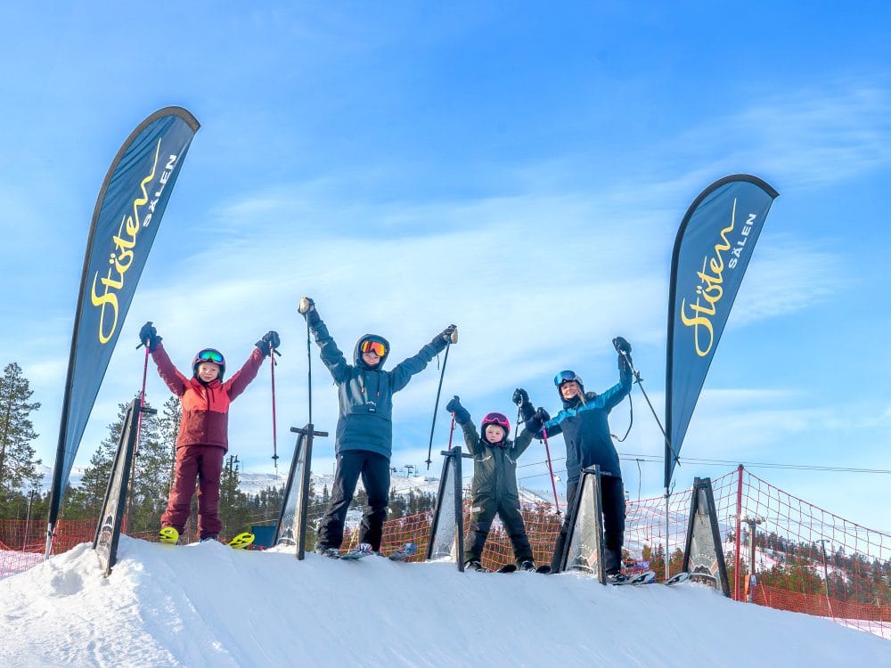 Fire personer på ski poserer med stave ved Stöten-flag i solrigt skiområde