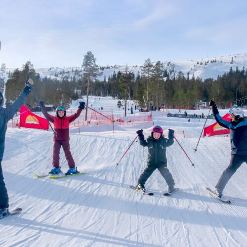 Fyra personer står på skidor och poserar med stavar i soligt skidområde med lift i bakgrunden