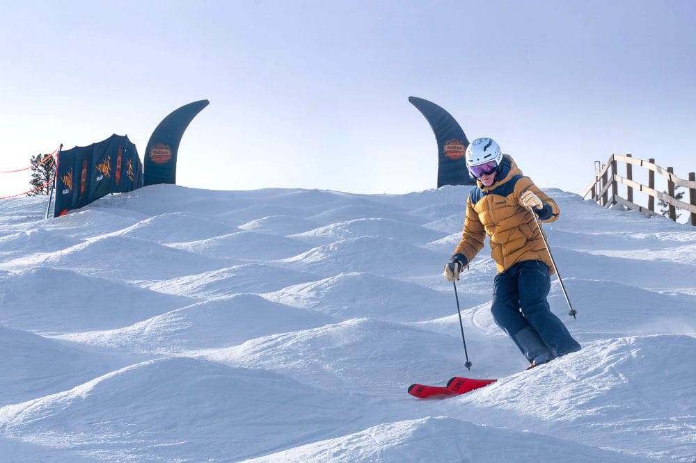 En person står på ski på pukler med sne og blå himmel i et solrigt skiområde