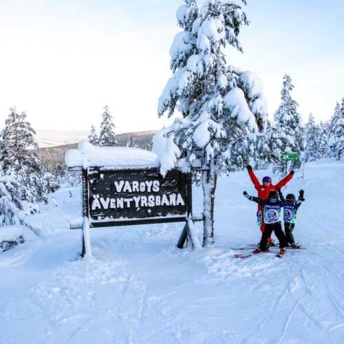 Tre personer på skidor poserar vid skylt som visar Vargys Äventyrsbana i snötäckt landskap