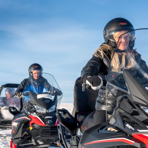 Two snowmobiles on mountain trail, woman leads family tour with children in sledge