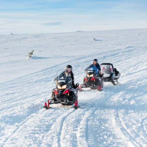Two snowmobiles with sleds drive down a mountain slope in a sunny winter landscape