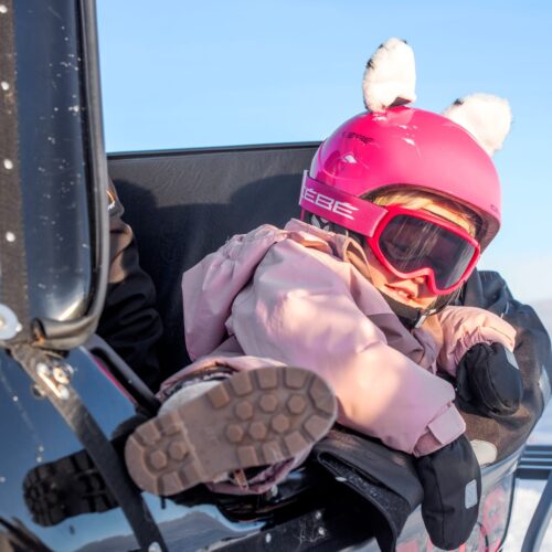 Children in sleds behind snowmobiles, pink helmets and winter clothes in a sunny mountain landscape
