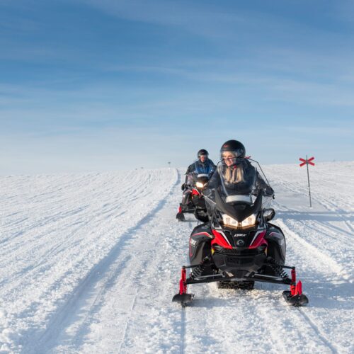 To personer kører snescooter på snedækket bjergsti under klar blå himmel