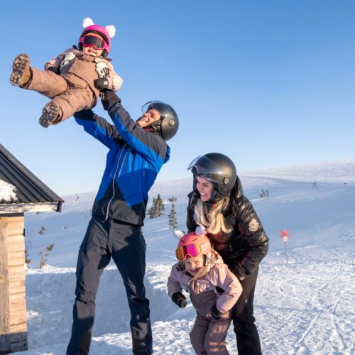 Familie på skibakke løfter barn i vintertøj, solrig dag med snedækket landskab