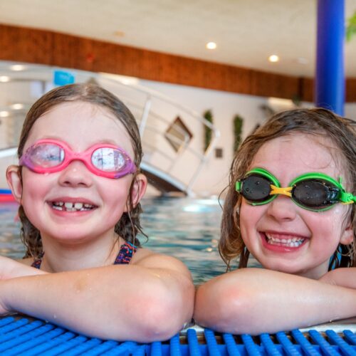 To glade børn med svømmebriller læner sig op ad bassinkanten indendørs.