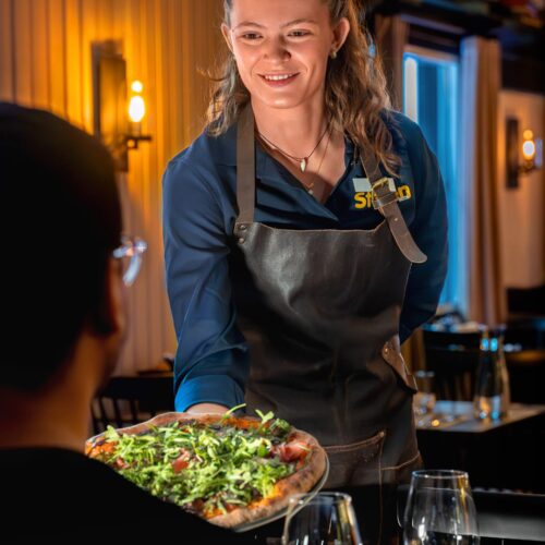 Waiters in leather aprons serve pizza with arugula and ham in a stylish restaurant setting.