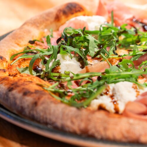 Nygräddad pizza med lufttorkad skinka, ruccola, mozzarella och balsamvinäger på mörk tallrik.