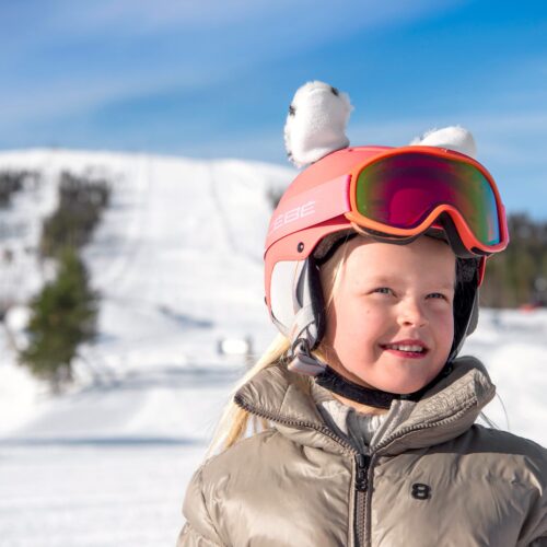 Person i beige skijakke og pink hjelm med ører står i solrigt skiområde