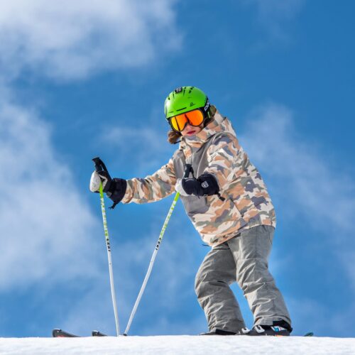 Person i grøn hjelm og camouflagejakke står på ski med stave under klar blå himmel