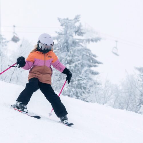 Barn åker skidor i snötäckt backe med rosa stavar och lift i bakgrunden