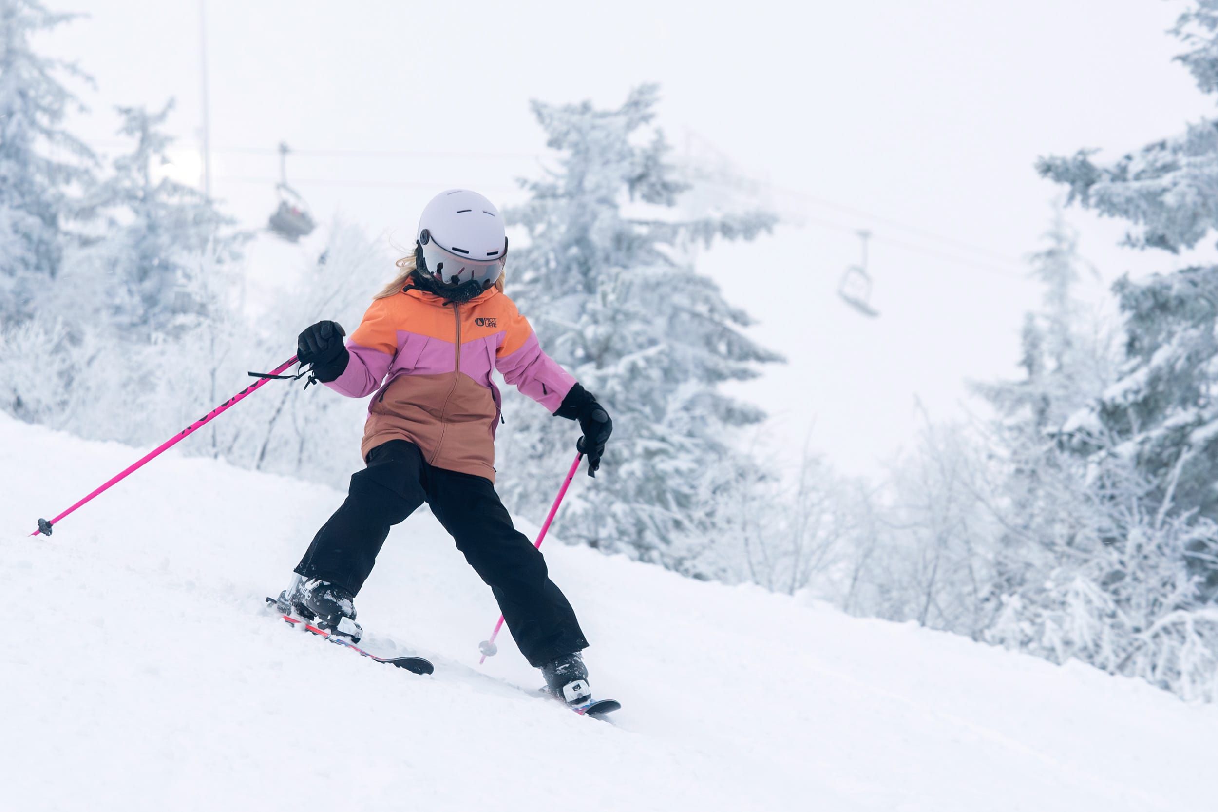 Barn åker skidor i snötäckt backe med rosa stavar och lift i bakgrunden