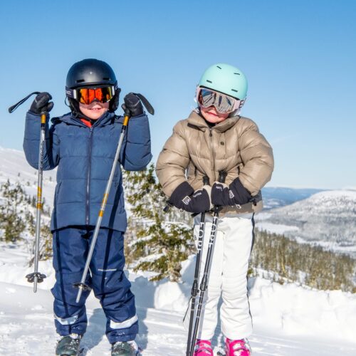 Två barn i skidkläder med hjälmar och stavar står i snöigt fjällandskap under klarblå himmel