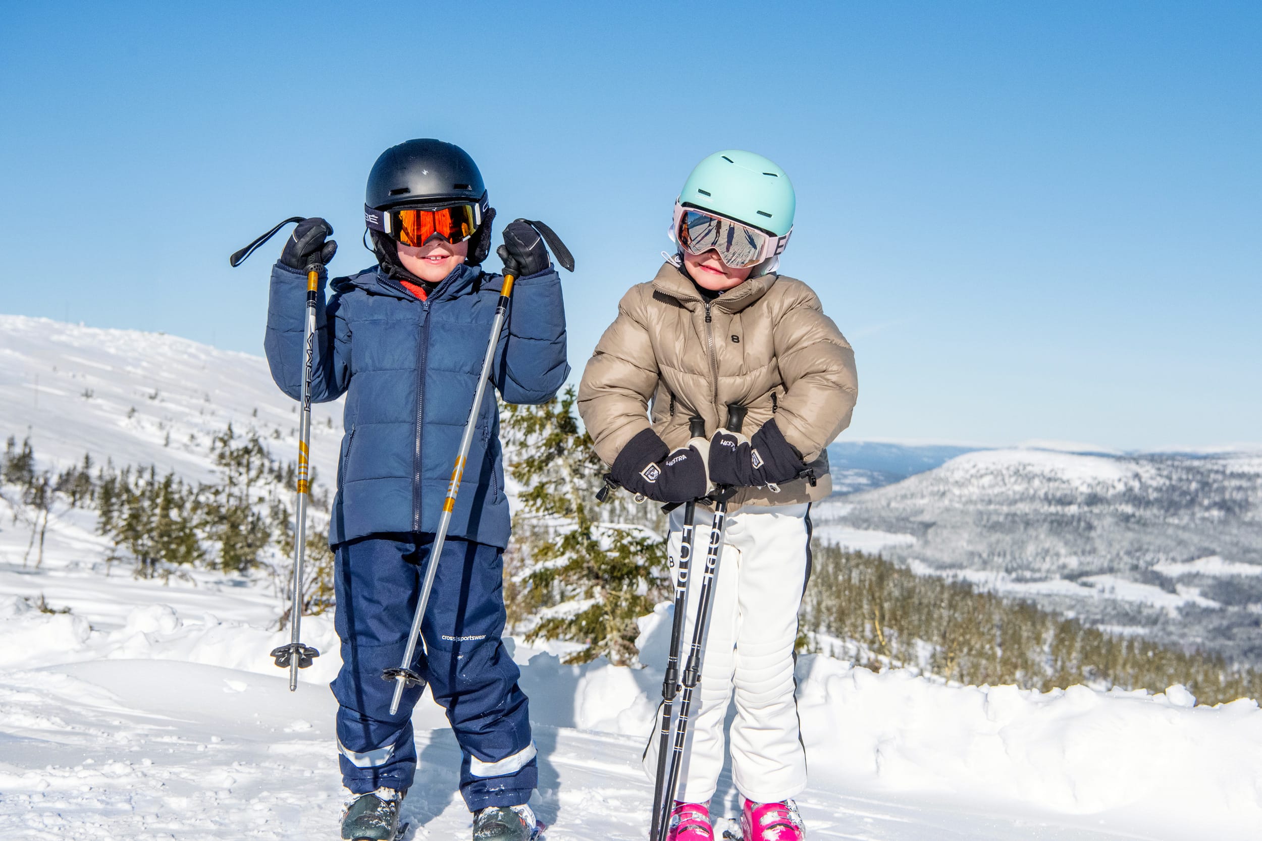To børn i skitøj med hjelme og stave står i et snedækket bjerglandskab under en klar blå himmel.