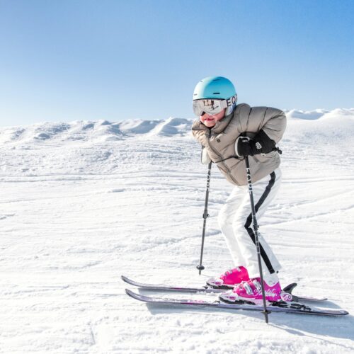 Barn åker skidor i solig backe med blå hjälm, rosa pjäxor och snölandskap i bakgrunden