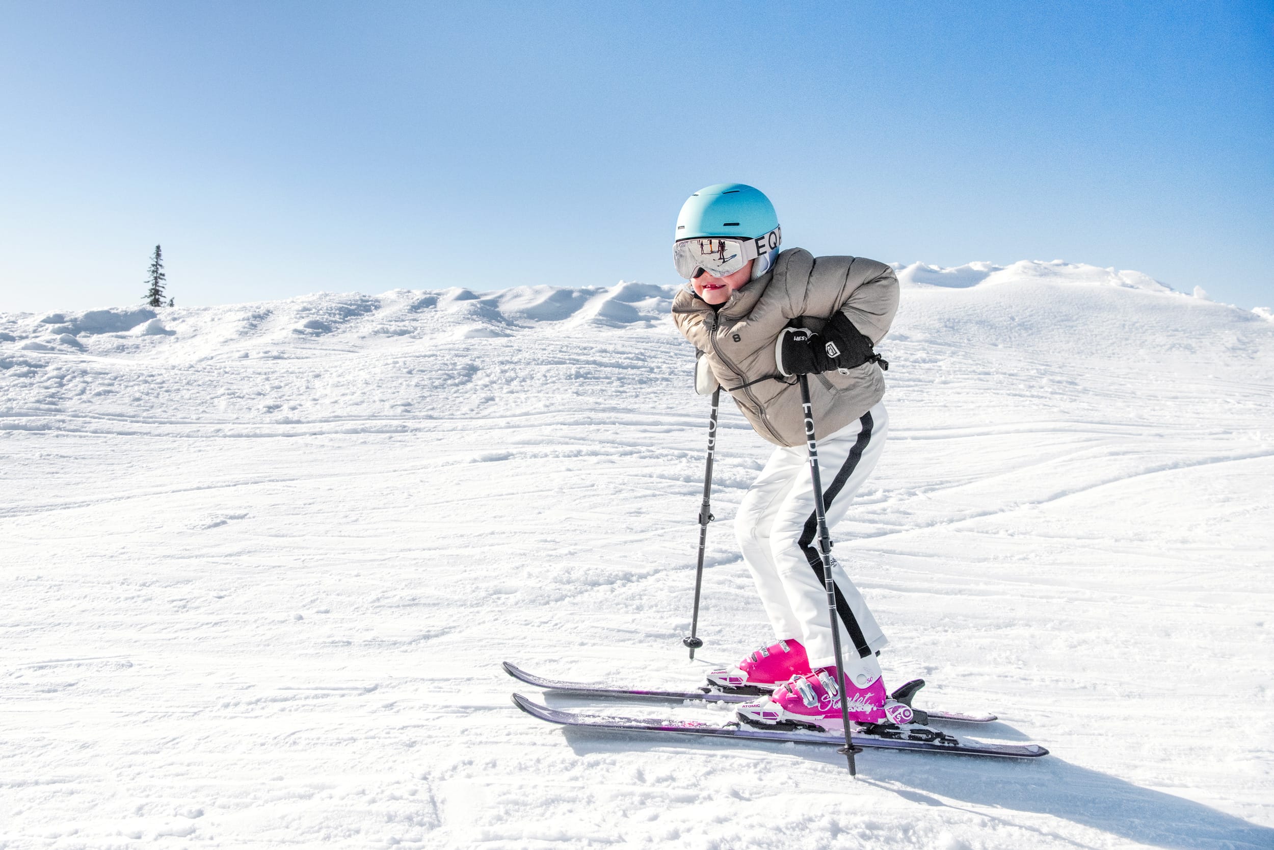 Barn åker skidor i solig backe med blå hjälm, rosa pjäxor och snölandskap i bakgrunden