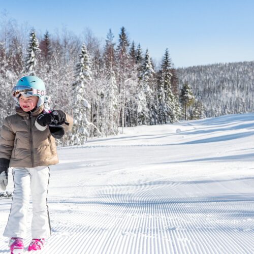 Barn i beige skidjacka och blå hjälm står på preparerad pist med snöklädda träd och klarblå himmel