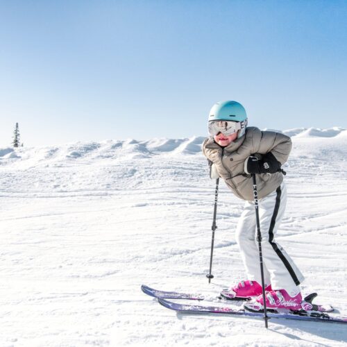 Børn står på ski på en solrig skråning med blå hjelme, lyserøde støvler og et snedækket landskab i baggrunden.