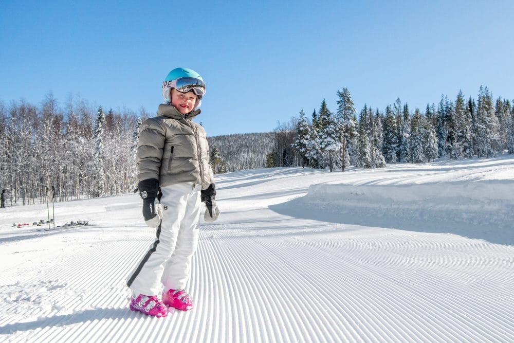 Children in beige ski jackets and blue helmets standing on a groomed piste with snow-covered trees and clear blue sky