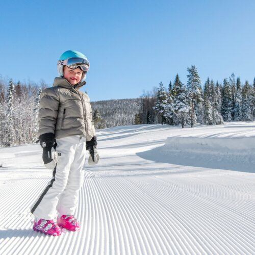 Barn i beige skidjacka och blå hjälm står på preparerad pist med snöklädda träd och klarblå himmel