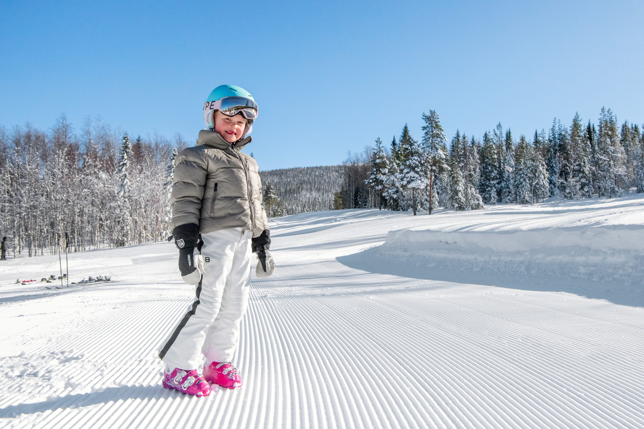 Barn i beige skidjacka och blå hjälm står på preparerad pist med snöklädda träd och klarblå himmel