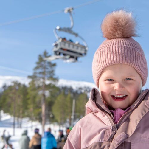 Person i lyserød skijakke og kasket står på solrig skibakke med stolelift og træer i baggrunden