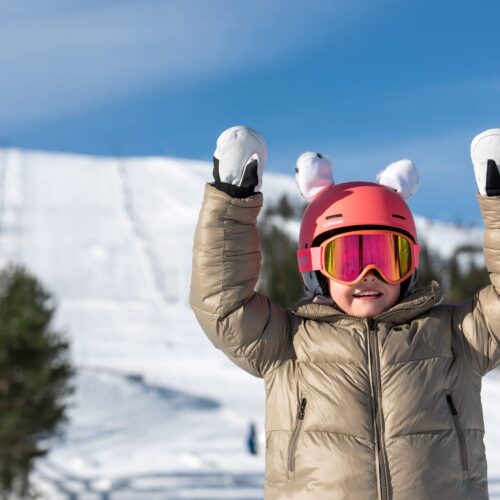 Barn i beige skidjacka med rosa hjälm och vita vantar står i solig skidbacke med snö och blå himmel
