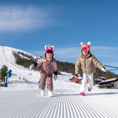 Två barn i skidkläder springer på preparerad pist med rosa hjälmar och enhörningsdetaljer under klarblå himmel