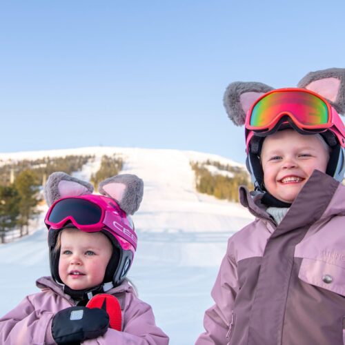 To personer i lyserødt skitøj og hjelme med ører står i et solrigt skiområde