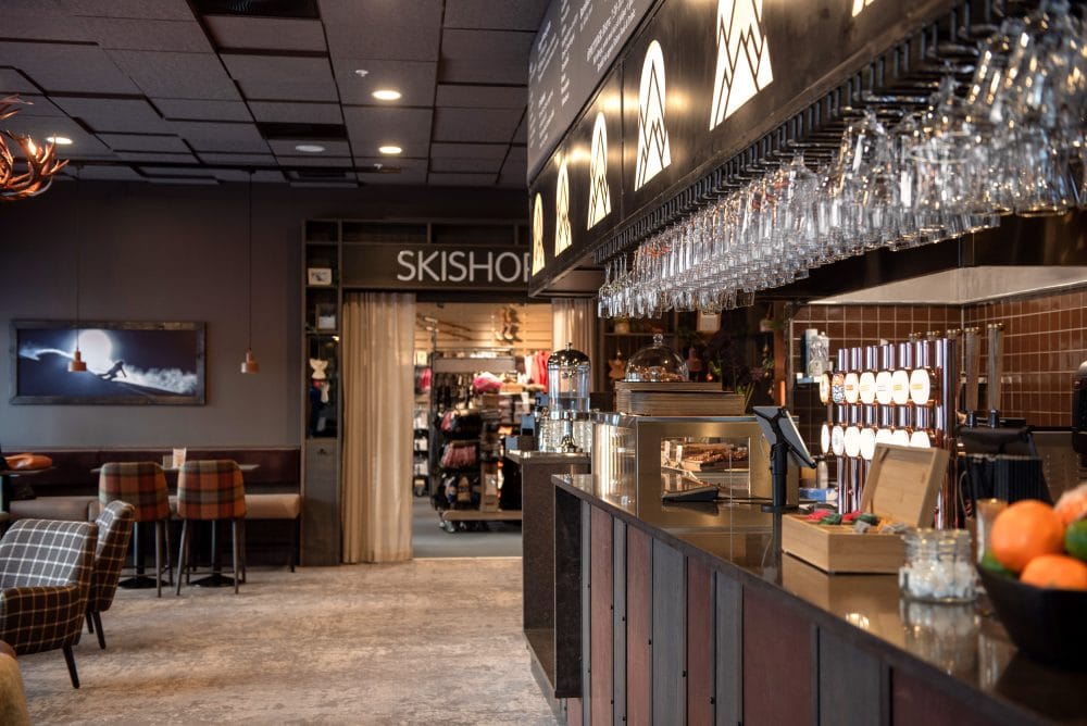 Modern bar med glas i takställ, meny ovanför och ingång till skidshop i bakgrunden.