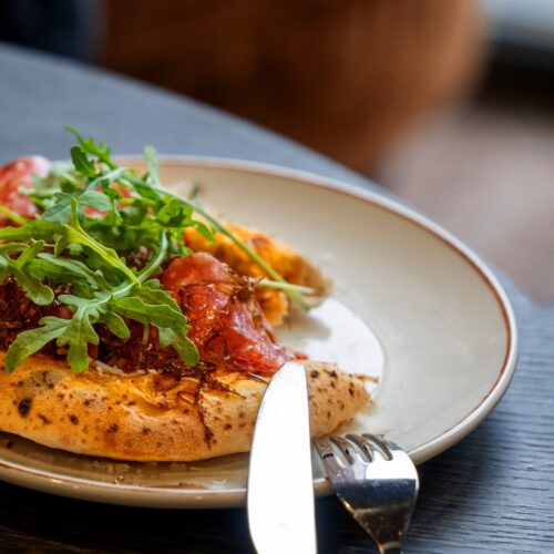 Pizza with air-dried ham, rocket and cheese on a light-coloured plate with cutlery on the side.