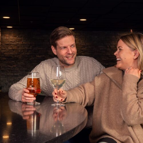 Two people at bar counter holding glasses of beer and white wine, copper coloured beer taps visible.
