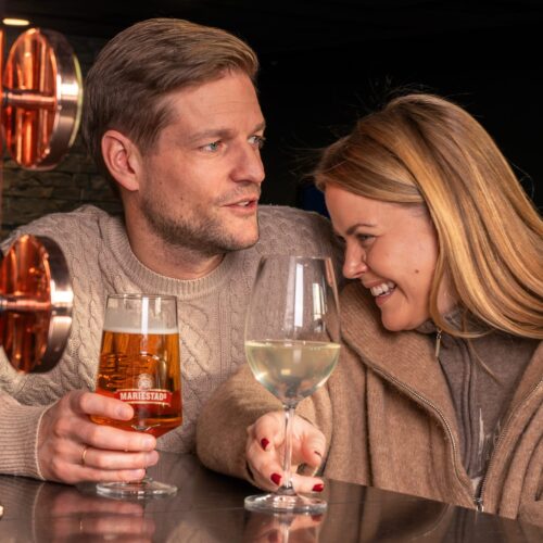 Two people at the bar counter hold glasses of beer and white wine next to copper-coloured beer taps.