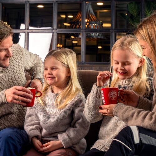 Four people are sitting in the lounge, holding red mugs with hot drinks at tables.