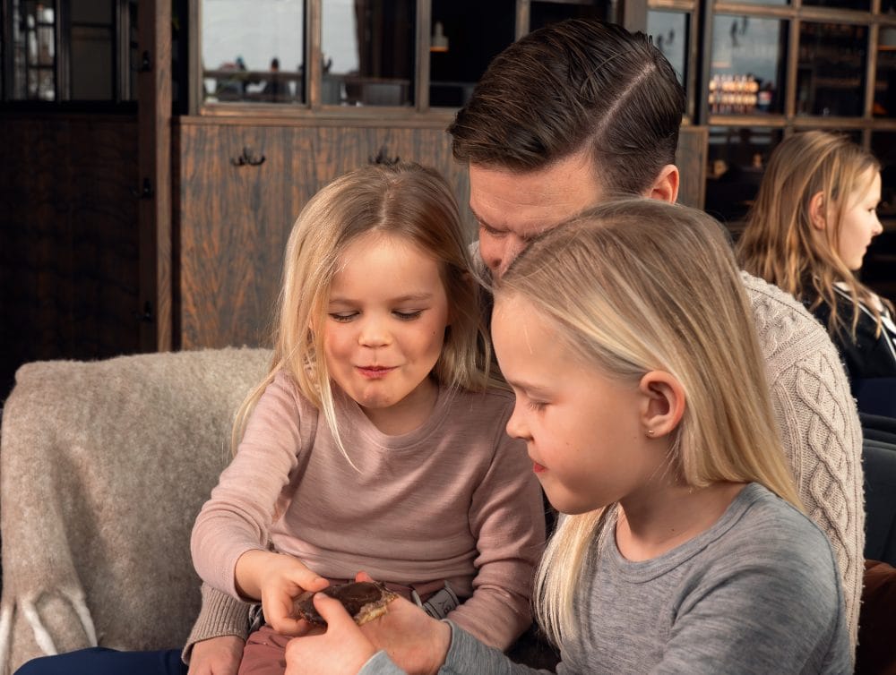 Två barn sitter nära vuxen och håller chokladbitar i mysig lounge med träinredning.