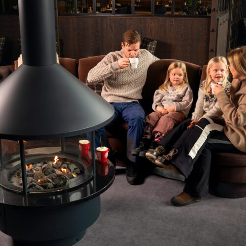 Fyra personer sitter vid rund soffa nära modern öppen spis och dricker varma drycker.