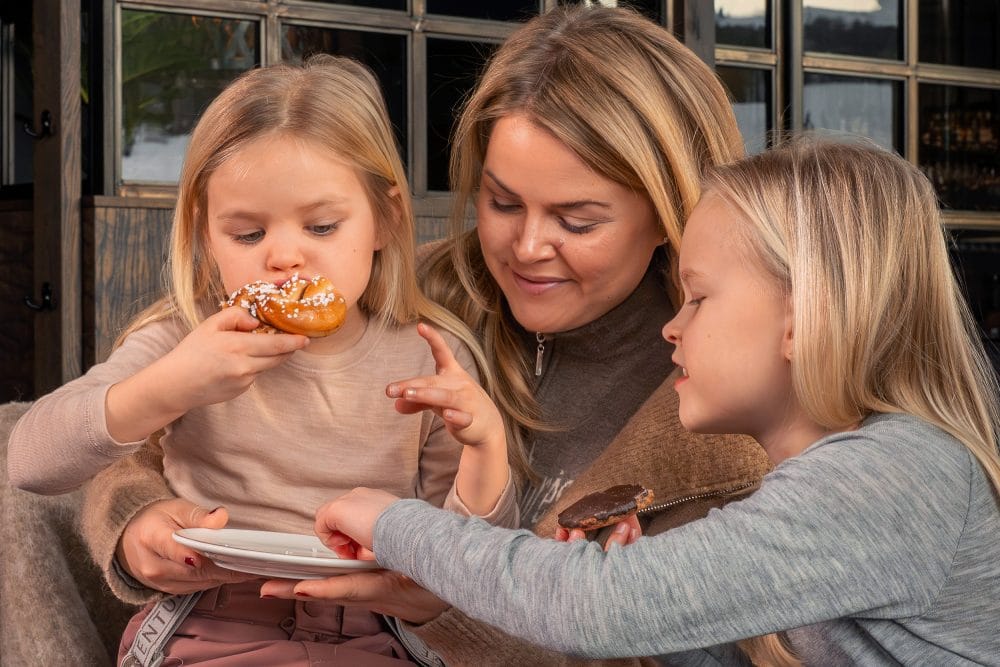 Tre personer sitter nära varandra, håller tallrik och chokladkaka i mysig lounge-miljö.