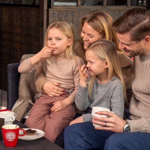 Four people sitting at table with hot drinks and cakes in cosy lounge near fireplace.