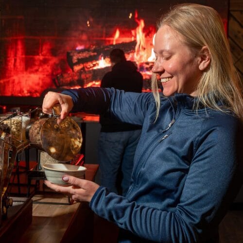 En person hælder müsli i en skål ved morgenmadsbuffeten med en stor pejs i baggrunden.