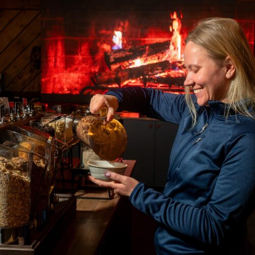 Person filling bowl with muesli at breakfast buffet, background with burning fireplace.