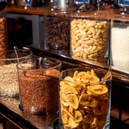 Glass containers with banana chips, nuts, raisins, coconut flakes and seeds on buffet tables.