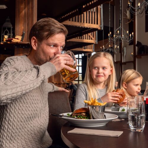 People sit at restaurant tables with burgers, fries and drinks in a rustic setting with wooden details.