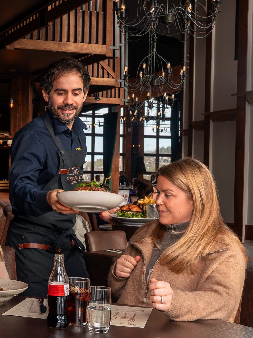 Serveringspersonal håller tallrik med mat vid dukat restaurangbord med drycker, glas och rustik inredning.