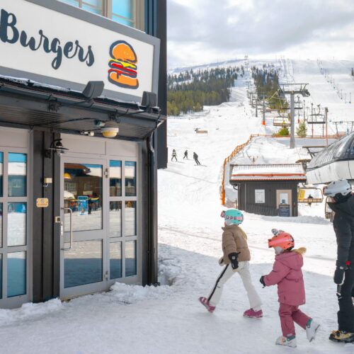 Restaurant sign Burgers at ski slope, people in helmet and ski clothes walking on snow covered ground.