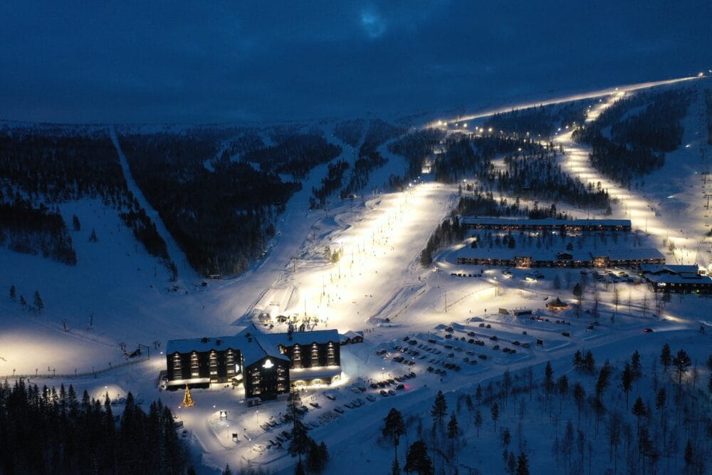 Upplyst skidområde nattetid med hotell, parkeringsplats och sammanhängande skidbackar i snölandskap.