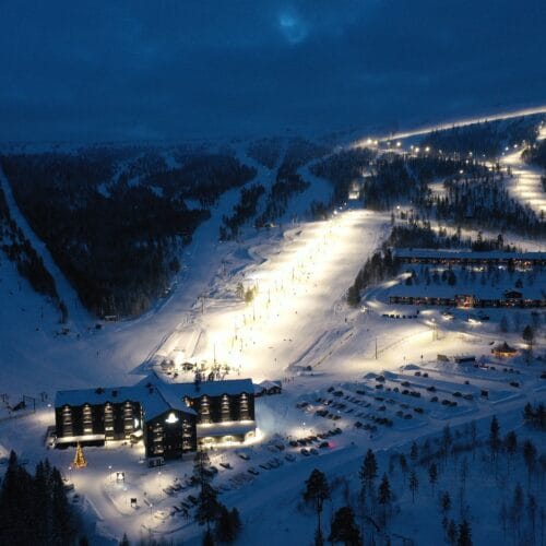 Upplyst skidområde nattetid med hotell, parkeringsplats och sammanhängande skidbackar i snölandskap.