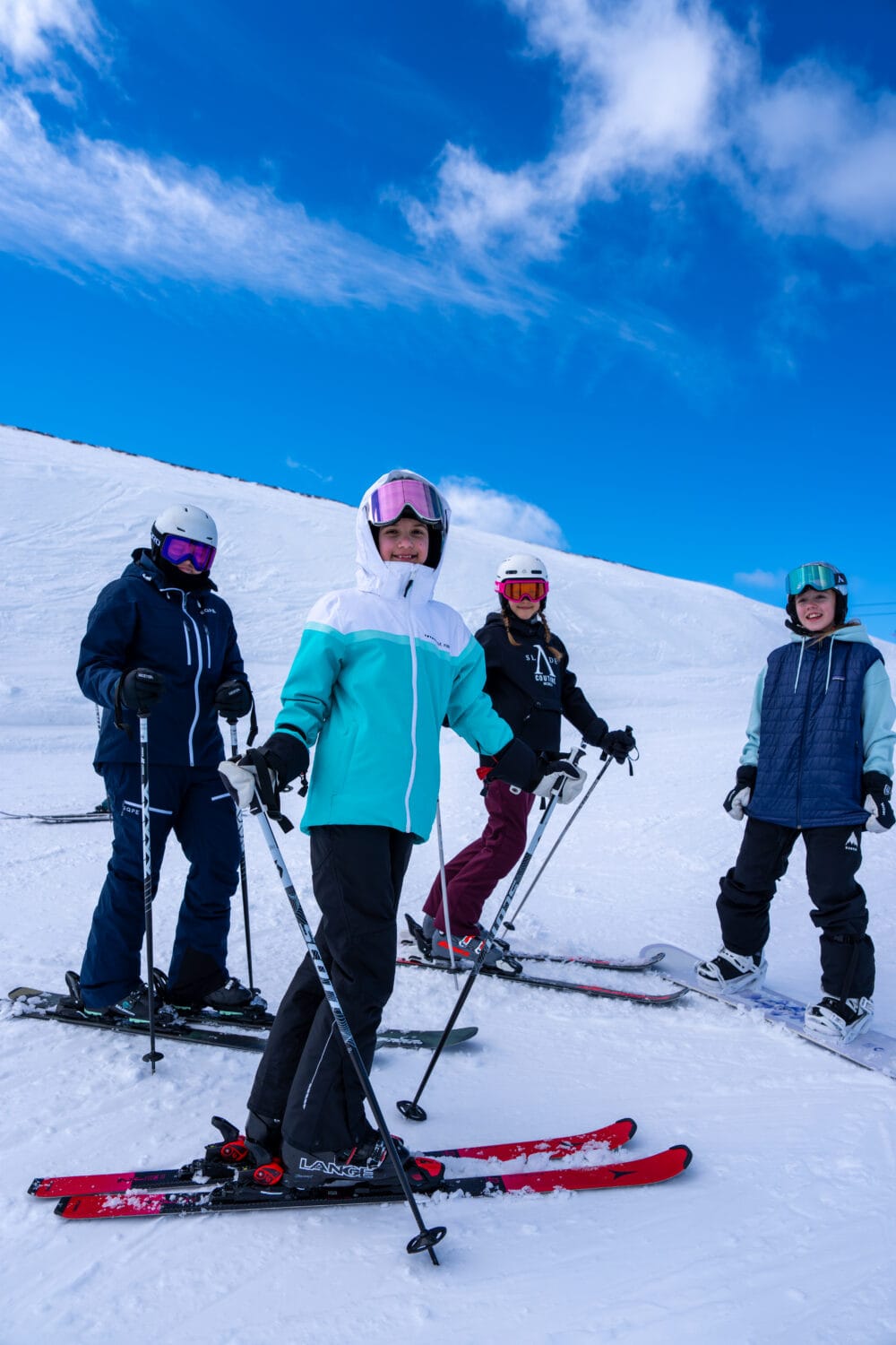 Four people standing on a ski slope with skis and poles under a clear blue sky and snow-covered landscape.