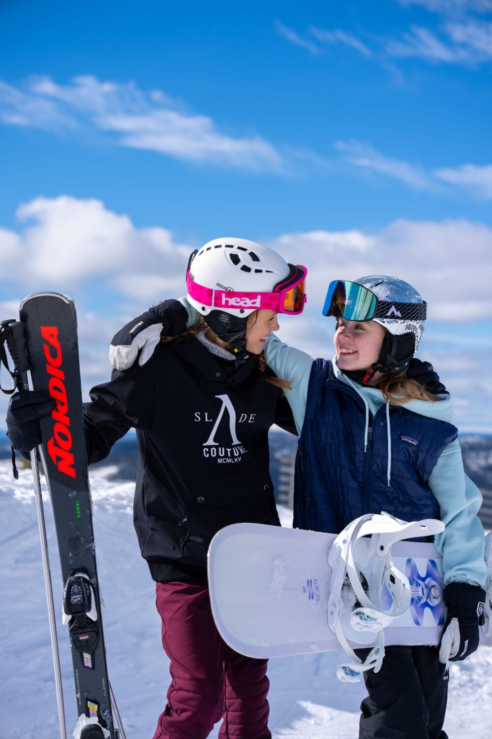 To mennesker på en skibakke med ski og snowboard, armene om hinanden, bjerge og blå himmel.