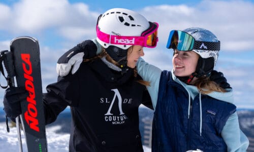 Two people on a ski slope with skis and snowboards, arms around each other, mountains and blue sky.